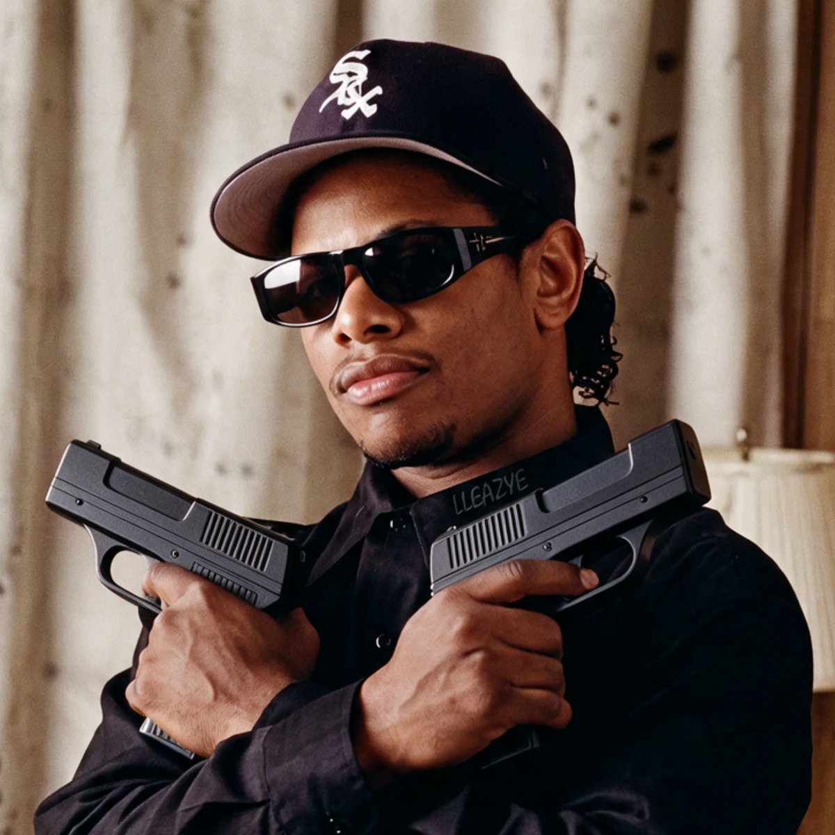A man wearing a black baseball cap with a white logo, black sunglasses, and a black button-up shirt holds two black semi-automatic pistols in a studio setting with a warm-toned curtain and wooden wall in the background.
